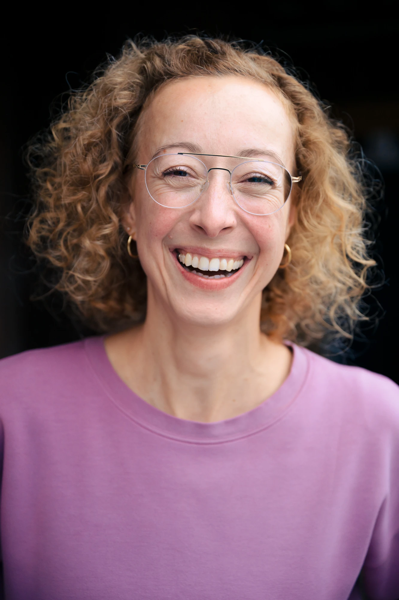 Herzlich lachendes Portrait von Heike Stier, Psychologische Beraterin, im fliederfarbenen Pullover vor dunklem Hintergrund